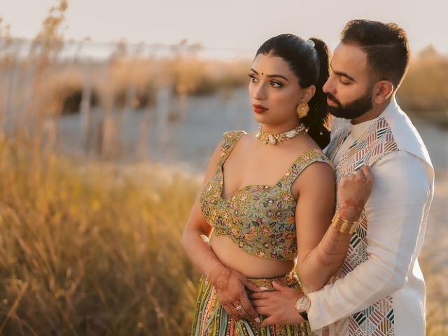 A couple stands in an embrace outdoors, with the woman wearing a colorful dress and the man in white attire, surrounded by tall grasses.