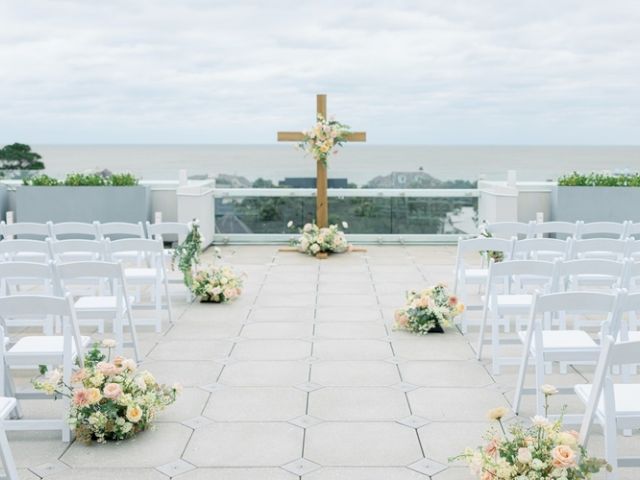 An outdoor wedding setup features white chairs, floral arrangements, and a wooden cross overlooking a scenic view with cloudy skies.