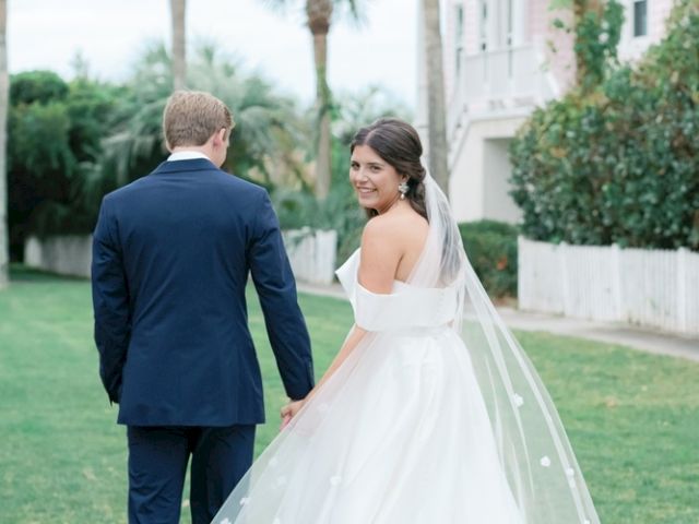 A bride and groom are walking hand in hand on a grassy path, with trees and a building in the background.
