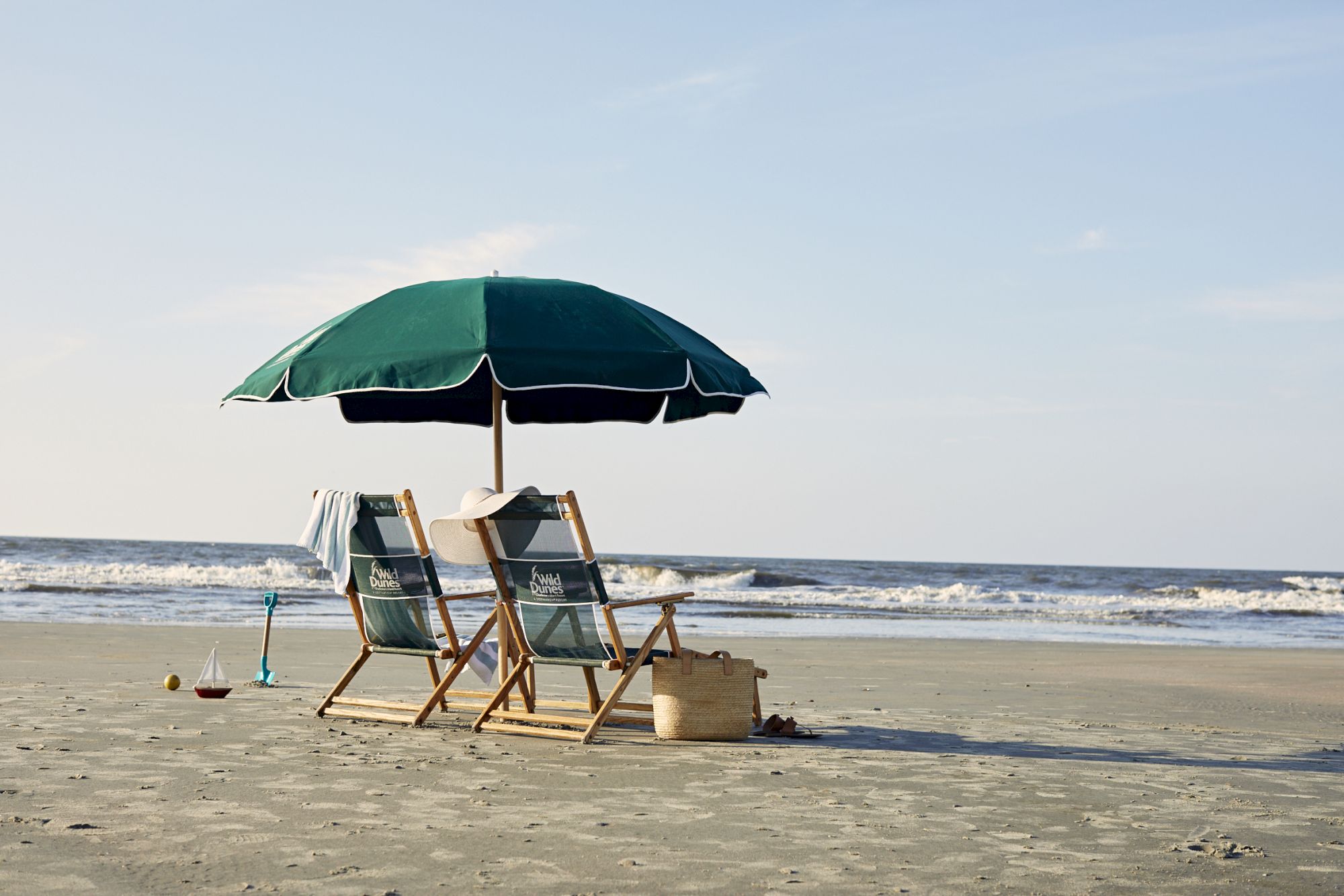 Two beach chairs and a green umbrella on sand by the sea, with waves and a few items like towels and a basket visible in the scene.