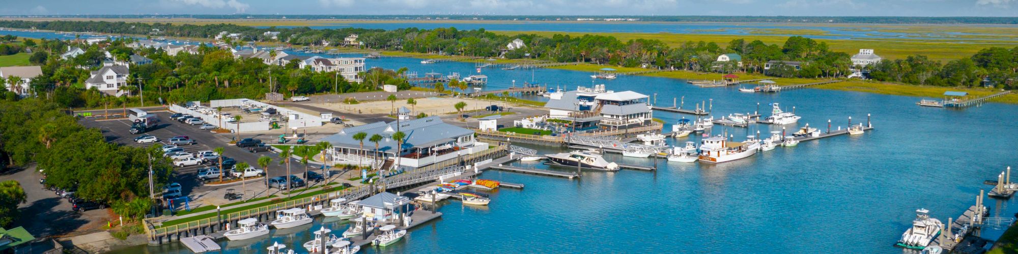 The image shows a marina with several boats docked, a parking area, and surrounding greenery under a blue sky with clouds.