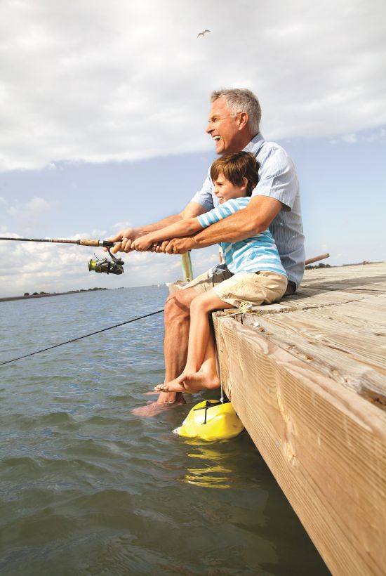 A person and a child are sitting on a dock fishing together, enjoying a peaceful moment by the water under a partly cloudy sky.