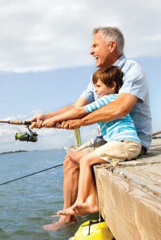 A man and child are fishing together, sitting on a wooden dock overlooking a body of water, both appear happy and relaxed.