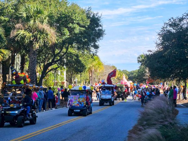 A lively street parade with colorful floats, people walking and riding golf carts, trees lining the road, and a bright blue sky.