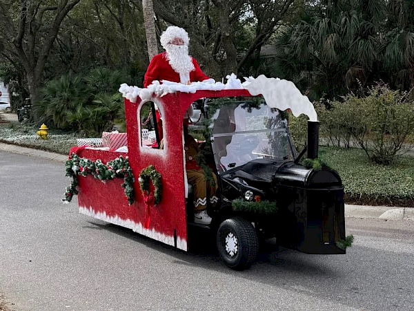 A person in a Santa suit is riding a small vehicle decorated like a holiday train, complete with festive garlands, on a road.