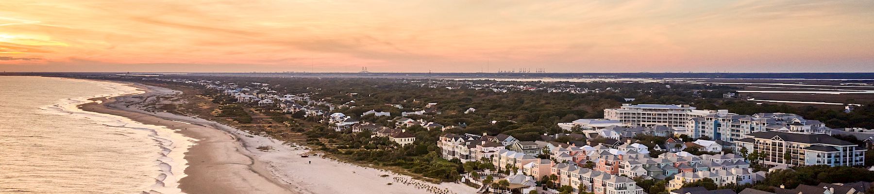 Aerial view of a coastal town with sandy beaches, buildings, and a calm sea under a colorful sunset sky.