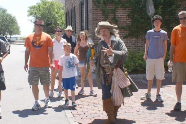 A person in historical attire leads a group of tourists on a sunny street, with brick buildings in the background.