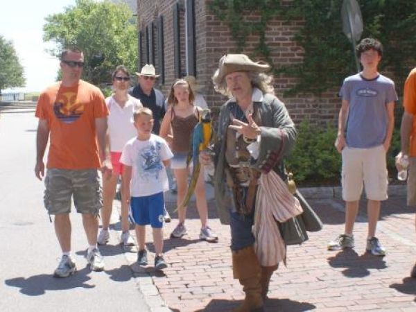 A person in historical attire leads a group of tourists on a sunny street, with brick buildings in the background.