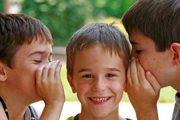 Three kids are in the image; two are whispering secrets to the smiling one in the middle.