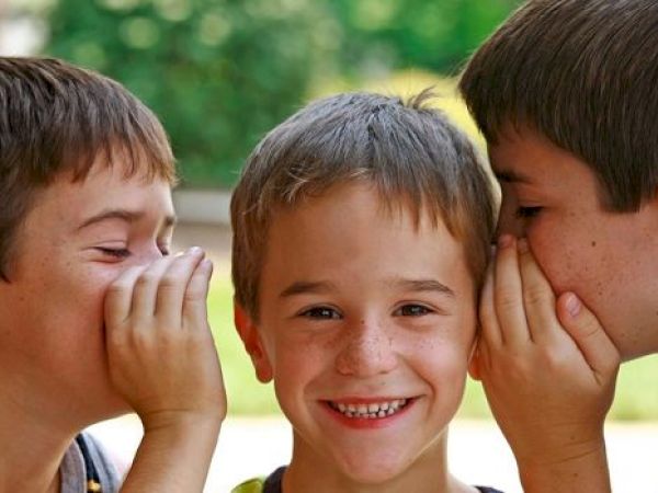 Three kids are in the image; two are whispering secrets to the smiling one in the middle.