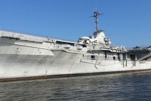 The image shows a large military aircraft carrier docked near a body of water under a clear blue sky.