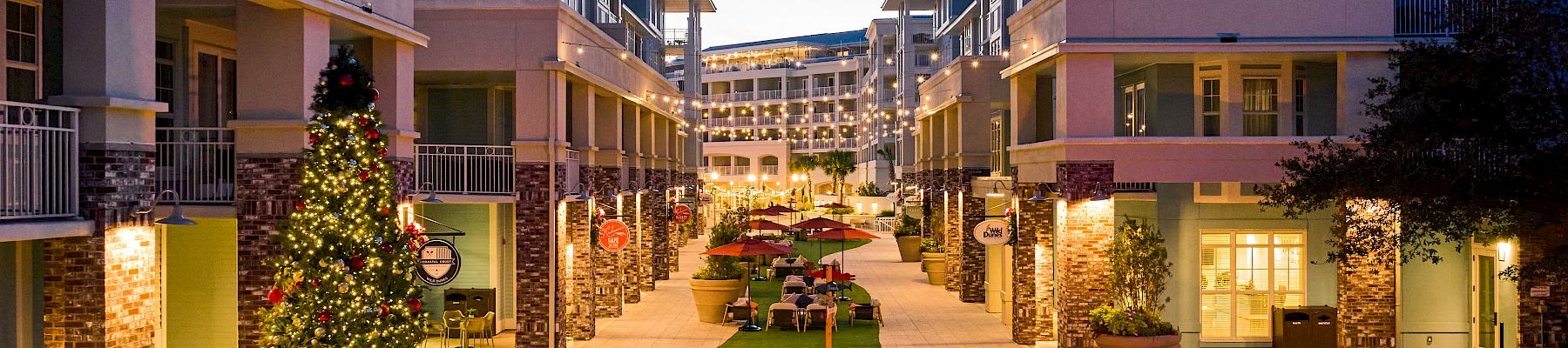The image shows a courtyard with a lit fire pit, surrounding chairs, and a Christmas tree, flanked by multi-story buildings at dusk.