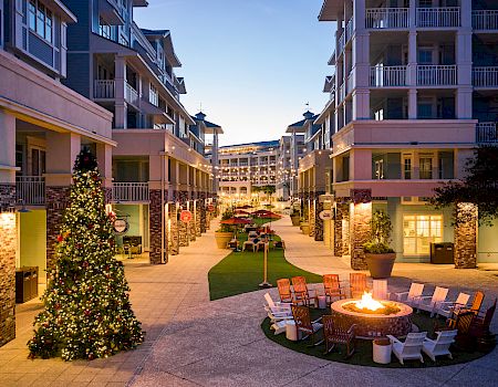 The image shows a courtyard with a lit fire pit, surrounding chairs, and a Christmas tree, flanked by multi-story buildings at dusk.