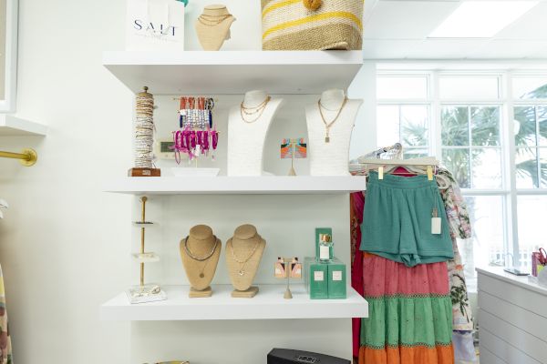 A boutique display with jewelry, a basket, bags, and a colorful dress on a rack in a bright, airy shop setting.