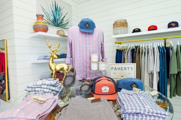 The image shows a retail display with shirts, caps, and accessories on a round table, surrounded by neatly hung clothes on racks in a bright room.