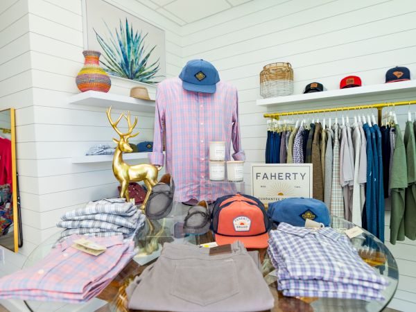 The image shows a retail display with shirts, caps, and accessories on a round table, surrounded by neatly hung clothes on racks in a bright room.