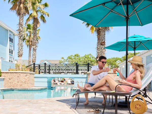A couple relaxes by a pool, sitting on lounge chairs under umbrellas, surrounded by palm trees and buildings in the background.