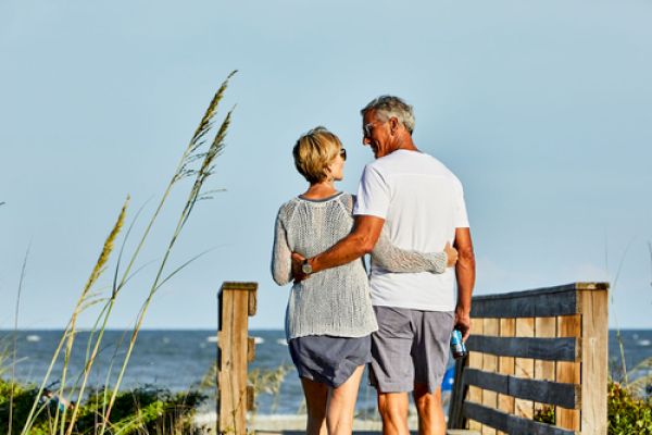 A couple walks arm in arm on a wooden path towards the beach, with grass and the ocean in the background.