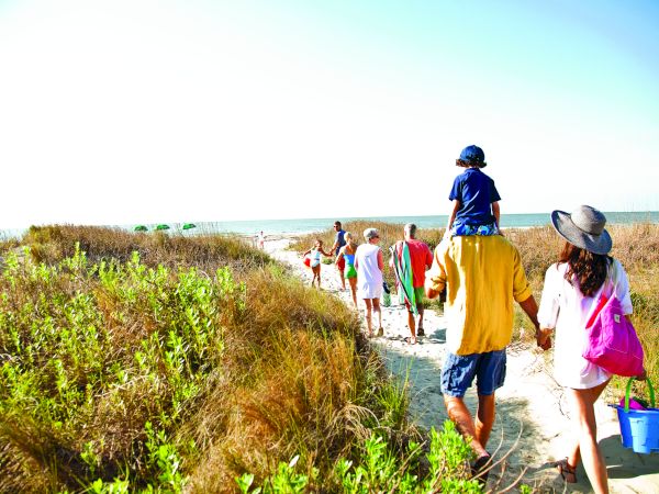 A group of people walking along a sandy path toward the beach, surrounded by vegetation, under a clear sky.