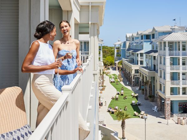 Two people are chatting on a sunny balcony overlooking a scenic street lined with modern buildings and greenery.