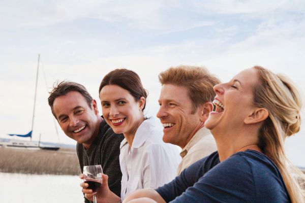 A group of four people are outdoors by the water, smiling and enjoying a relaxed moment together with a sailboat in the background.