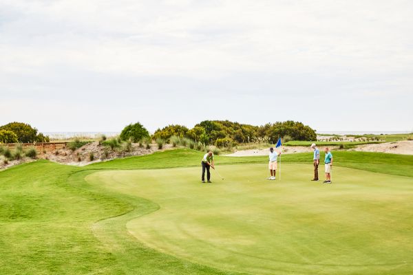 A group of people playing golf on a lush green course, with one individual taking a putt while others watch, under a cloudy sky.