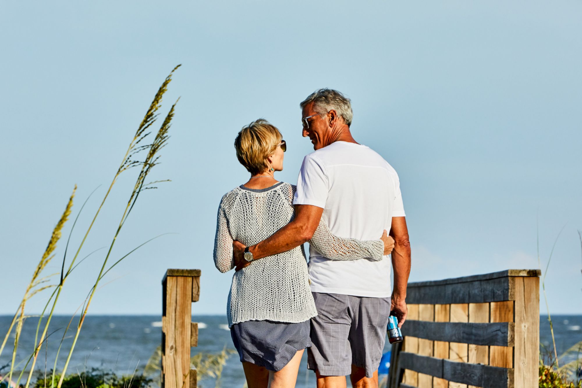 An older couple is walking arm in arm on a wooden pathway by the sea, enjoying a peaceful moment with the ocean in the background.