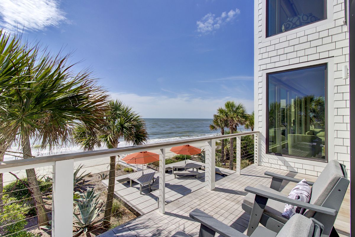 A beachside balcony with chairs overlooks the ocean, surrounded by palm trees and a clear blue sky.