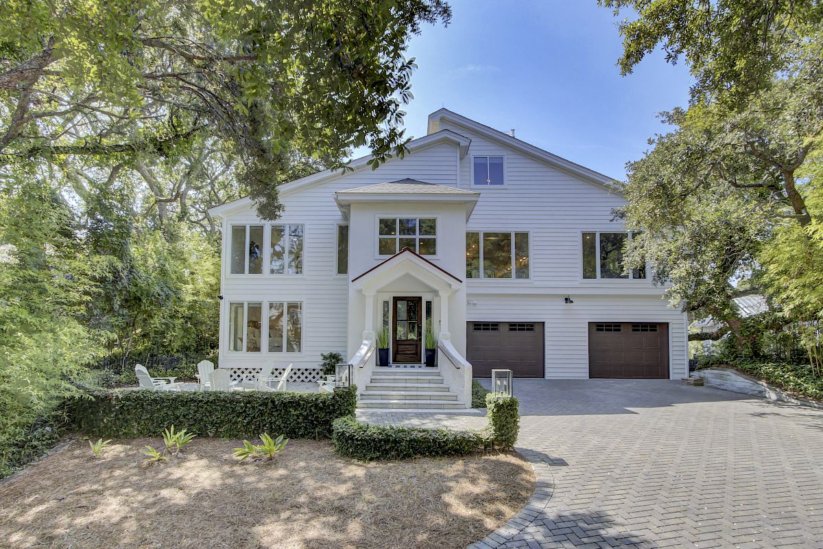 A large, white two-story house with multiple windows, a front porch, and a three-car garage, surrounded by trees and greenery.
