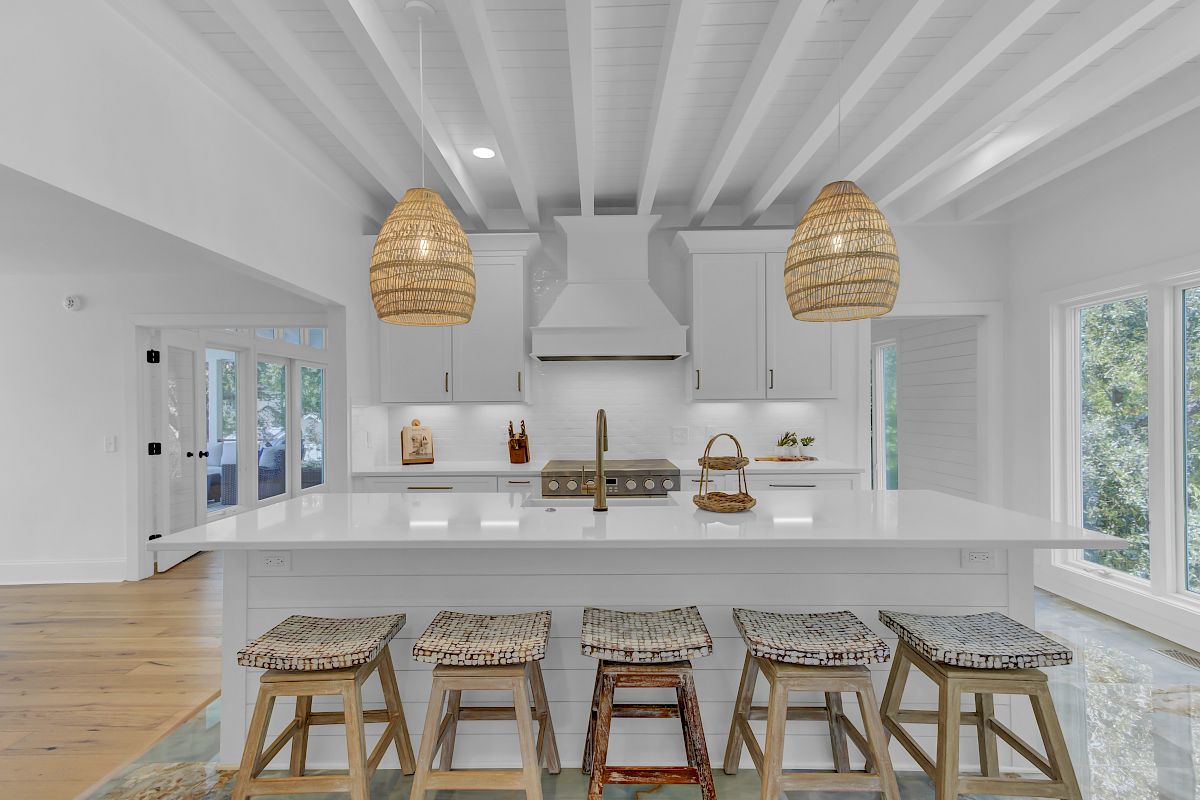 A bright kitchen with a white island, four stools, pendant lights, and wooden accents, featuring a range hood and decorative items.