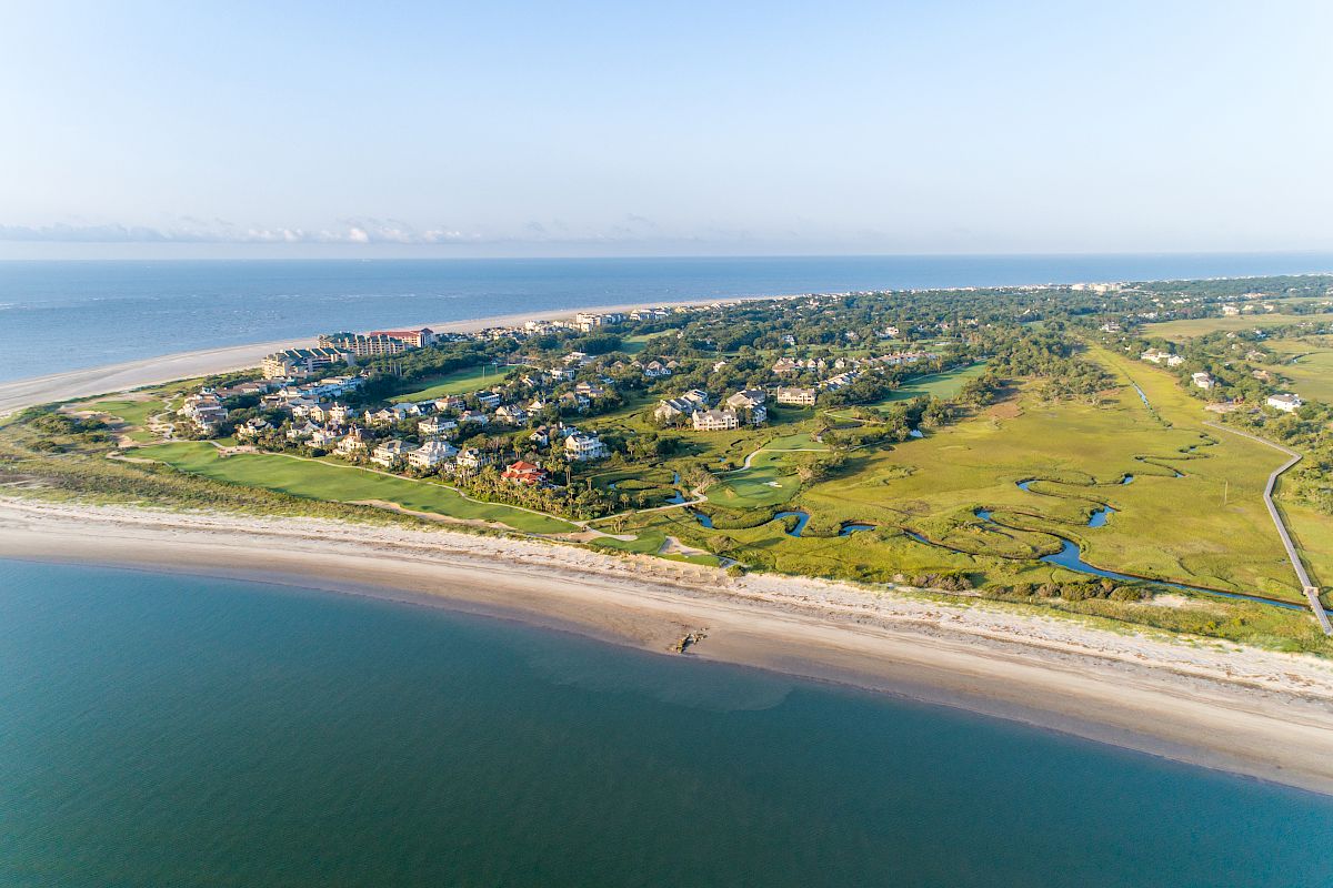 Aerial view of a coastal area with a beach, houses, greenery, and water, showcasing a serene and picturesque landscape.