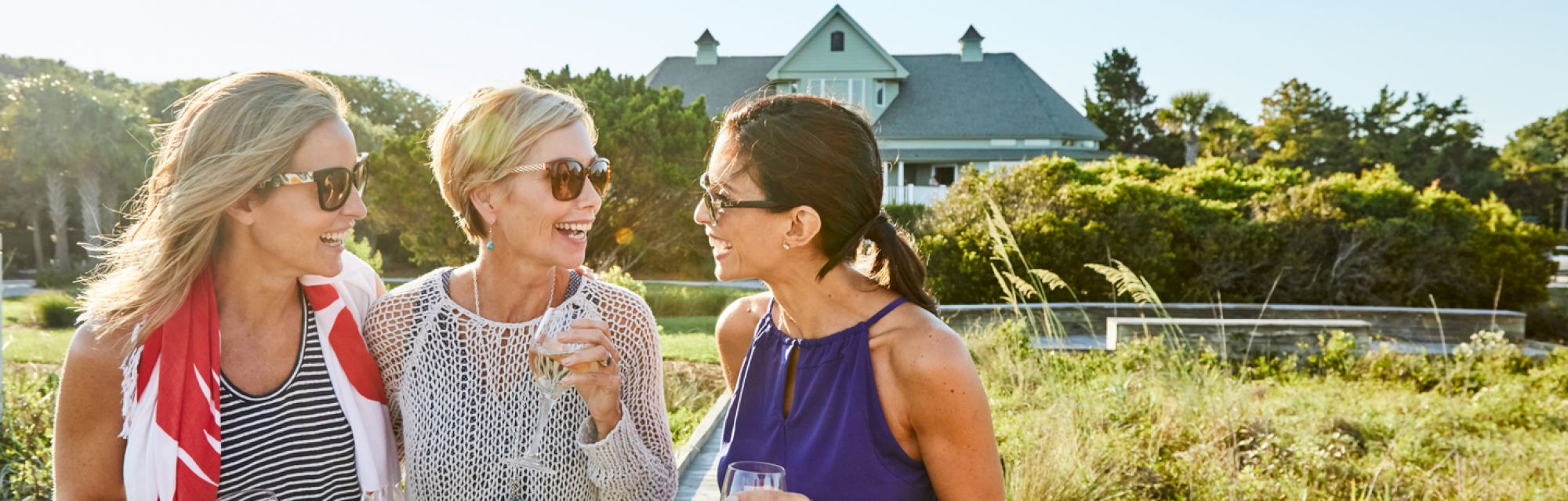 Three women smiling and holding drinks outdoors, with a large house and greenery in the background.