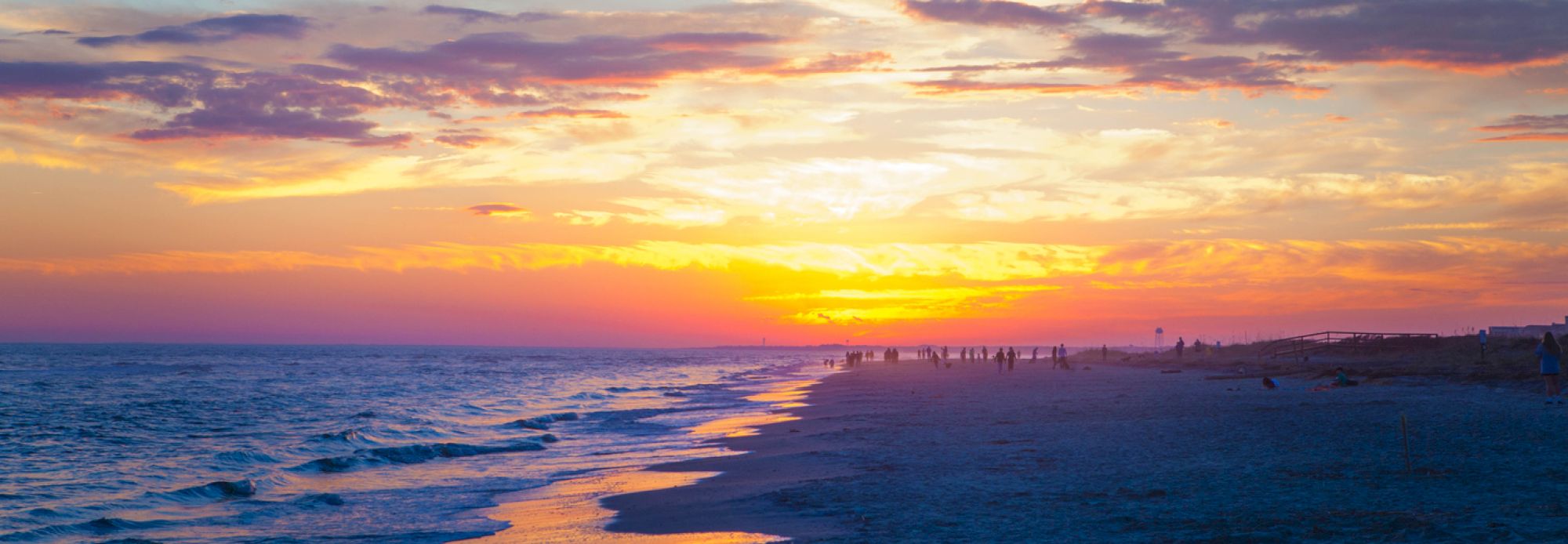 A beach at sunset with colorful skies and gentle waves, silhouettes of people walking along the shoreline in the distance.