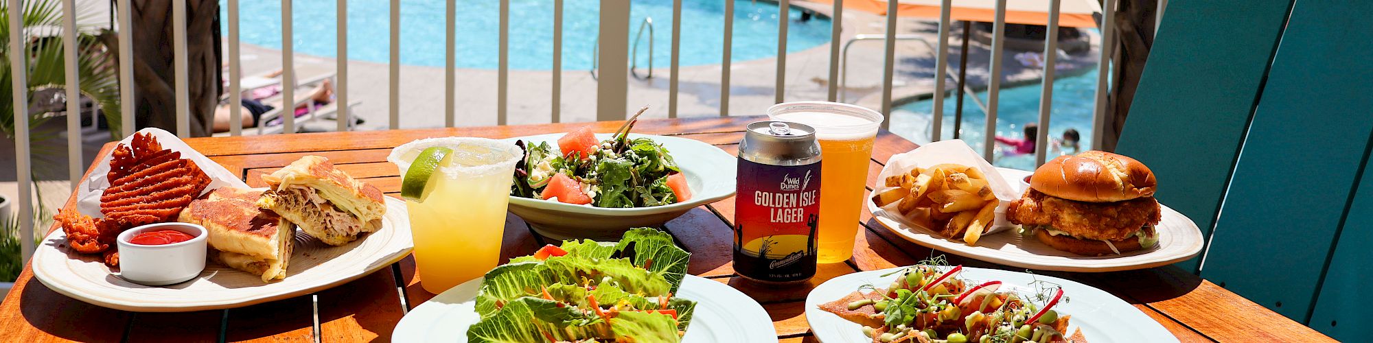 A poolside table with diverse dishes: salads, sandwiches, a drink, and a pool view in the background.