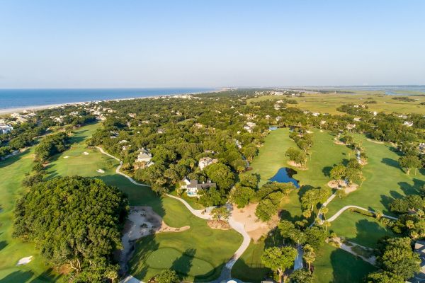 Aerial view of a golf course with surrounding greenery, residential areas, and the ocean in the background under a clear blue sky.