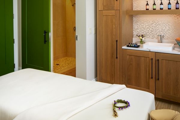 The image shows a serene spa room with a massage table, fresh towels, a shower area, and a counter with bottles and a sink for treatments.