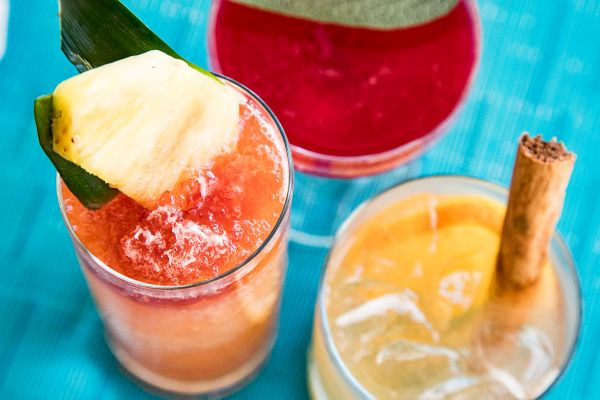 Three colorful cocktails on a blue surface, garnished with pineapple, a cinnamon stick, and fruit slices.