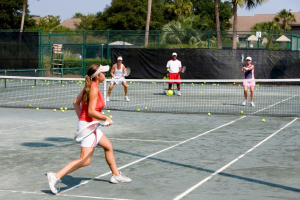 People are playing tennis on a court surrounded by scattered balls, with a green fence and vegetation in the background.