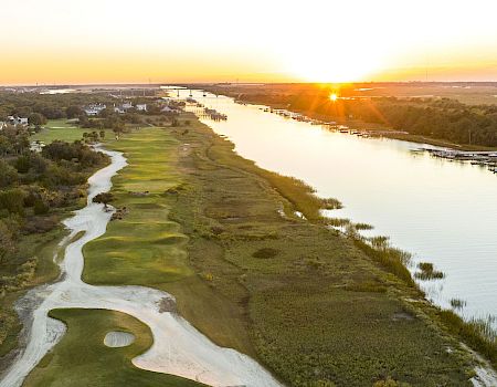 The image shows a scenic view of a golf course by a river during sunset, with lush greenery and a winding path.