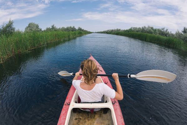 A person in a red kayak paddling through a narrow waterway, surrounded by green reeds and under a partly cloudy sky.