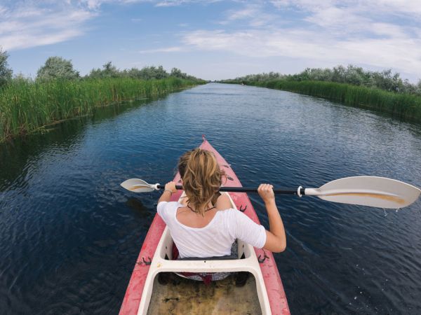 A person in a red kayak paddling through a narrow waterway, surrounded by green reeds and under a partly cloudy sky.