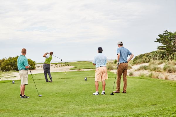 Four people are on a golf course; one is swinging a club while the other three watch, surrounded by greenery and a clear sky.