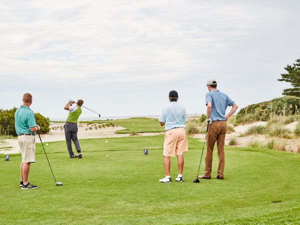 Four people are on a golf course; one is swinging a club while the other three watch, surrounded by greenery and a clear sky.