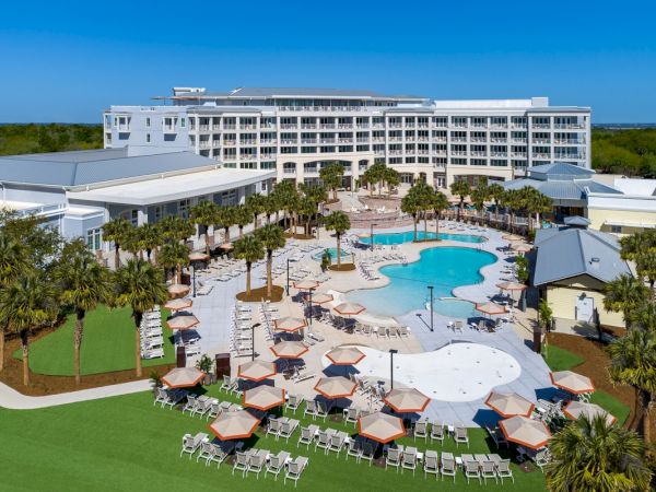 The image shows a resort with a large pool, surrounded by lounge chairs, umbrellas, and palm trees, adjacent to a multi-story building.
