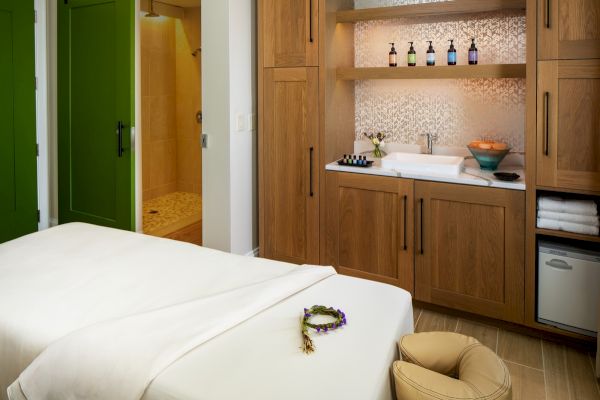 A serene spa room with a massage table, towels, lotions, a sink, and green doors leading to a shower area. A calming atmosphere is present.
