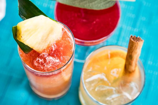 Three colorful cocktails on a blue surface: one with a pineapple garnish, one red, and one with an orange slice and cinnamon stick.