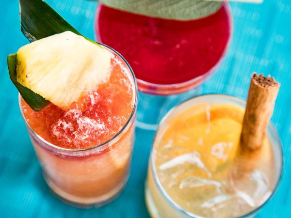 Three colorful cocktails on a blue surface: one with a pineapple garnish, one red, and one with an orange slice and cinnamon stick.