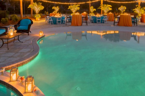 A poolside event setup with tables, chairs, string lights, and lanterns. The ambiance is warm and inviting, perfect for an evening gathering.
