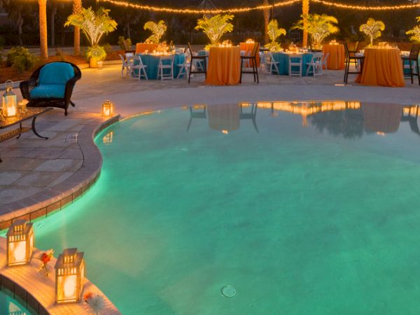 A poolside event setup with tables, chairs, string lights, and lanterns. The ambiance is warm and inviting, perfect for an evening gathering.