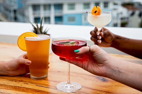 Three hands hold different drinks: a beer with an orange slice, a red cocktail, and a white drink with garnish, on a wooden table.